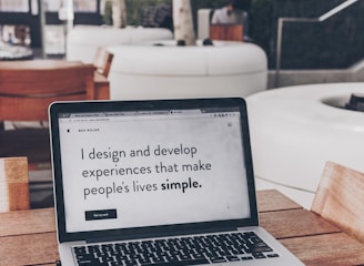 A laptop displaying a webpage with the text 'I design and develop experiences that make people's lives simple.' is placed on a wooden table. It is set in an outdoor seating area with white furniture and greenery in the background. The setting appears to be calm and conducive to work or relaxation.