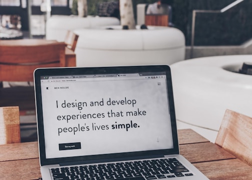 A laptop displaying a webpage with the text 'I design and develop experiences that make people's lives simple.' is placed on a wooden table. It is set in an outdoor seating area with white furniture and greenery in the background. The setting appears to be calm and conducive to work or relaxation.