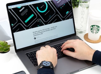 A person is typing on a MacBook Pro laptop surrounded by a smartphone, a green plant, a bottle of VOSS water, and a Starbucks paper cup. The laptop screen displays a UI/UX design with several circular graphic elements and a heading related to an iOS concept. The individual is wearing a smartwatch on their wrist.