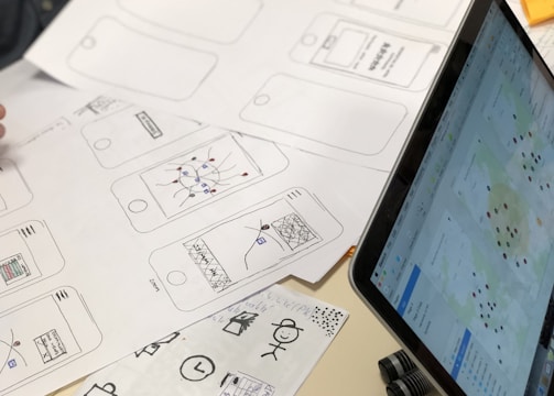 A series of drawn wireframes and sketches on white paper showcasing mobile app interface designs. The designs are accompanied by a laptop displaying a digital map with various data points. Pens and other sketching tools are partially visible, indicating a workspace focused on digital design and mapping.